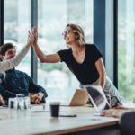 two women team members high-five in a business meeting room with two male colleagues