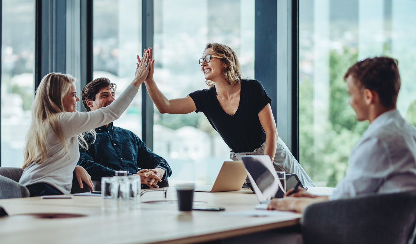 two women team members high-five in a business meeting room with two male colleagues